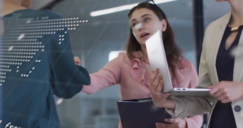Businesswomen reaching across glass wall, shaking hands while holding laptop and clipboard