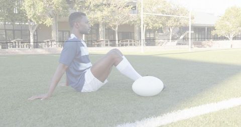 Male rugby player sitting on turf holding ball resting near goalposts on sunny field