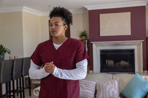 Home healthcare clinician standing with clasped hands in living room wearing maroon scrubs