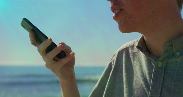 Young man holding a smartphone near his mouth, recording a voice message while enjoying a sunny day at the beach. Perfect for content related to mobile communication, outdoor technology use, vacation activities, beachfront lifestyles, or apps for voice messaging and virtual communication.