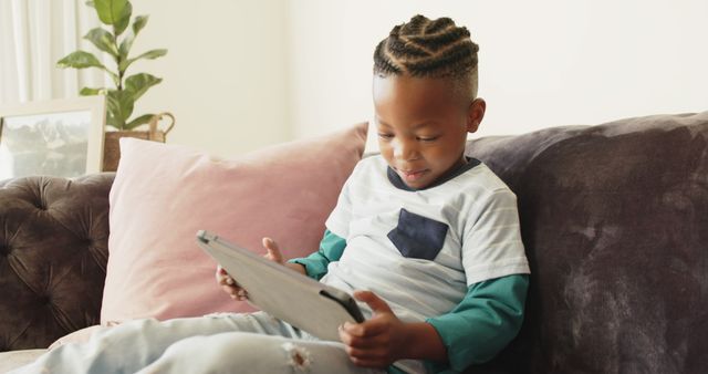 Young boy comfortably seated on a sofa, engrossed in using a tablet. Ideal for topics related to children's use of technology, screen time, modern parenting, or educational content for kids.