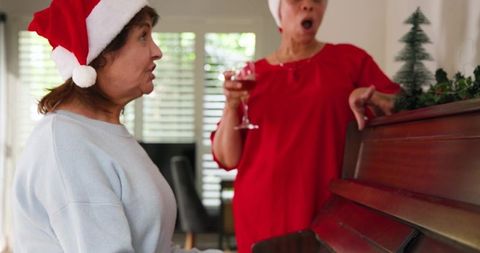 Mature women wearing santa hats singing duet at upright piano holding wine glasses