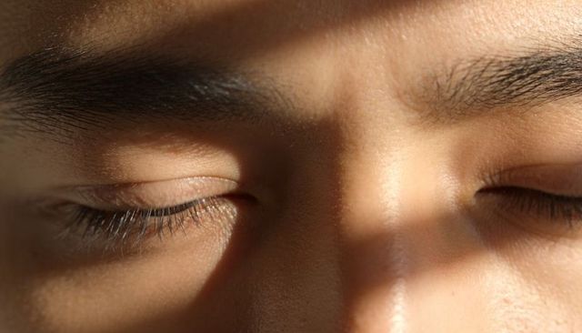 Sunlit closed eyes on male-presenting face with detailed skin texture and soft shadow band