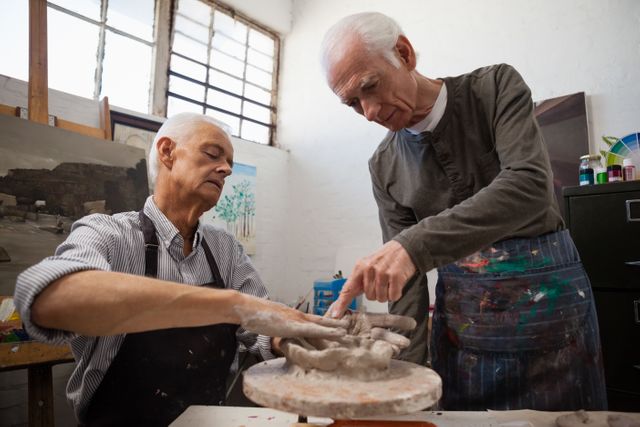 Two senior men are deeply engaged in pottery making in an art class. One man is assisting the other, guiding his hands as they work with clay. This image is perfect for illustrating themes of creativity, lifelong learning, teamwork, and the importance of artistic activities for seniors. It can be used in articles about senior education, art therapy, or community workshops.