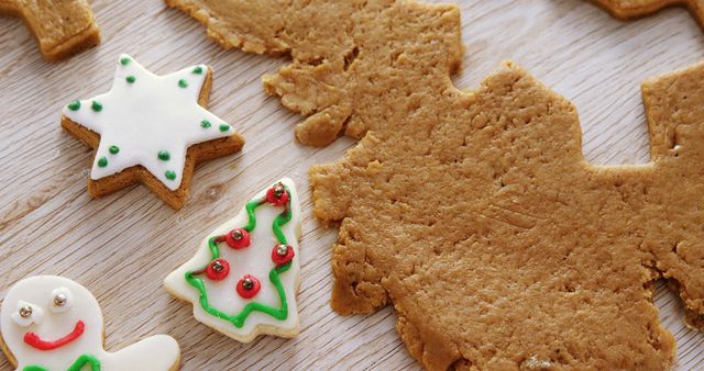 Decorated gingerbread cookies in festive shapes lie next to a sheet of cookie dough on a wooden surface, with copy space. These treats are often associated with holiday celebrations and home baking traditions.