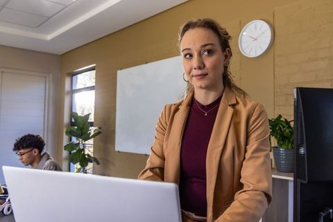 Professional woman standing at laptop in coworking office with whiteboard and clock