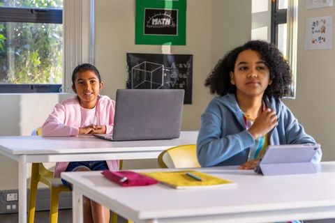 Preteen girls using laptop and tablet in sunlit classroom, blended learning and collaboration