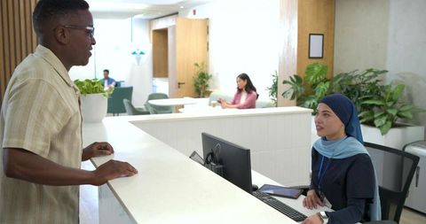 Guest leaning on reception desk while receptionist in blue hijab checking computer