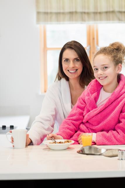 Mother and daughter are having breakfast together in their home kitchen. Both are wearing bathrobes, suggesting a relaxed and cozy morning at home. The mother is holding a mug, while the daughter is enjoying a bowl of cereal with a glass of orange juice. Perfect for use in family-focused marketing, advertisements promoting healthy lifestyles, or content highlighting family bonds and morning routines.