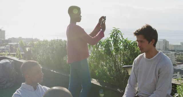 Group of friends socializing on a rooftop with cityscape in background. One person capturing moment with phone while others engage in conversation. Perfect for designs focused on social interactions, urban lifestyle, and outdoor activities.