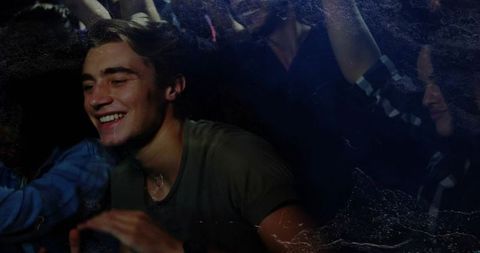 Young man smiling leaning forward in crowded club with raised arms and concert lights