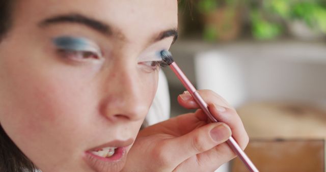 Image depicts woman applying blue eyeshadow using a makeup brush. Perfect for blogs, articles, or websites related to beauty, makeup tutorials, cosmetics products, and self-care routines. Useful for beauty brands' advertising campaigns or social media content promoting makeup tips.