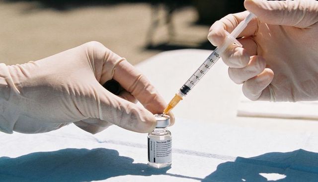 Gloved hands drawing vaccine into syringe from vial outdoors closeup with sunlit shadows