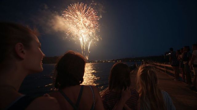 Women watching fireworks over lake from wooden pier at night, reflections and silhouettes