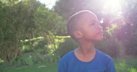 African american boy standing in sunlit garden looking up with thoughtful expression