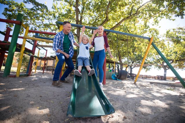 Parents enjoying time with their son at a playground. The boy is sliding down a slide while his parents watch and smile. Ideal for use in advertisements, family-oriented content, parenting blogs, and promotional materials for parks or recreational activities.