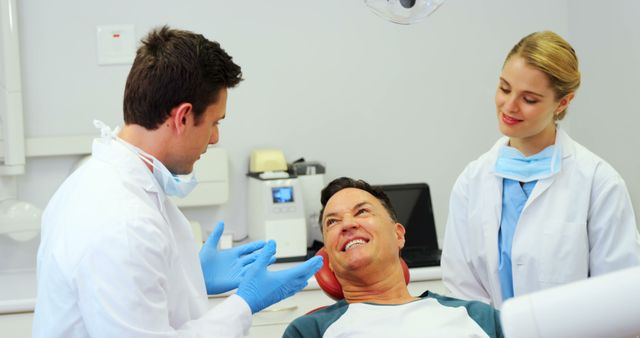 Dentist wearing gloves and lab coat providing a consultation to a patient sitting in a dental chair. Female dental assistant standing beside them, observing the interaction. Ideal for use in content related to dental health, medical services, healthcare advice, patient care, and promoting dental clinics.