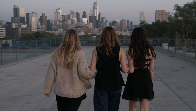 Three friends walking toward city skyline at dusk, linking arms on rooftop terrace