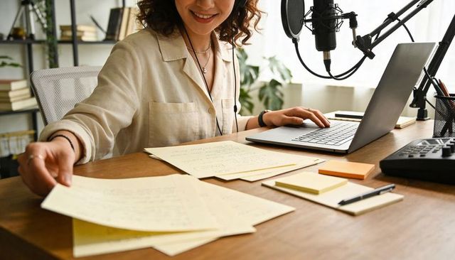 Woman recording podcast in home studio wearing headset reaching for notes