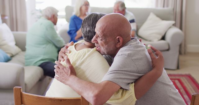 This stock photo displays a heartfelt moment where two senior friends are embracing warmly in a cozy living room with other seniors engaging in the background. This image can be used to depict themes such as friendship, elderly lifestyle, retirement community, and emotional connections. It is ideal for use in advertisements, websites, articles related to senior living, healthcare, or retirement homes.