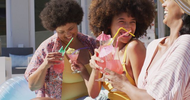 Three happy women share laughter while enjoying vibrant pink beverages during a sunny pool party. Ideal for vacation promotions, summer events, and lifestyle content focused on fun and relaxation.