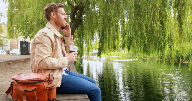 Young man talking on phone holding coffee by lake under willow tree - Download Free Stock Images Pikwizard.com