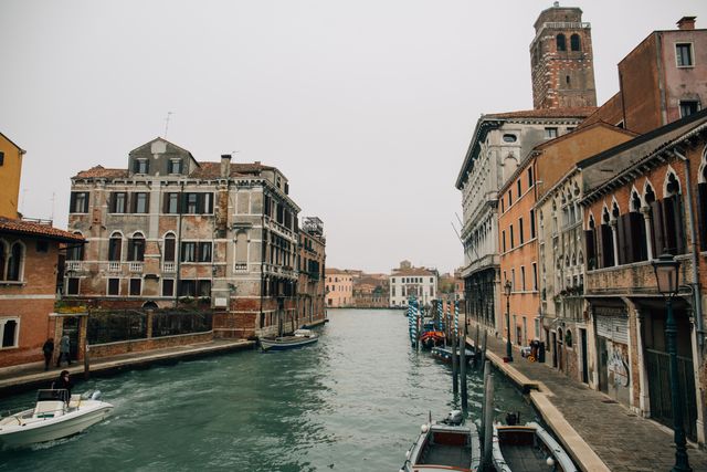 Capturing the timeless beauty of a canal in Venice, Italy, this image showcases historic buildings lining the waterway, with boats docked along the sides. The overcast sky adds a serene and moody ambiance. Ideal for travel blogs, tourism websites, architecture magazines, or as wall art highlighting Europe's rich cultural heritage.