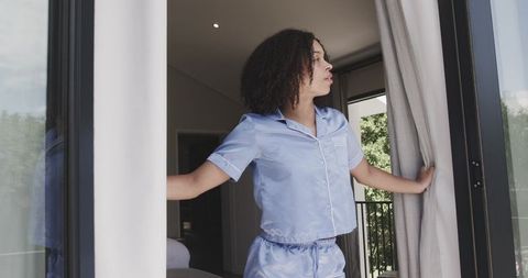 African american woman pulling curtains to balcony in blue satin pajamas morning routine