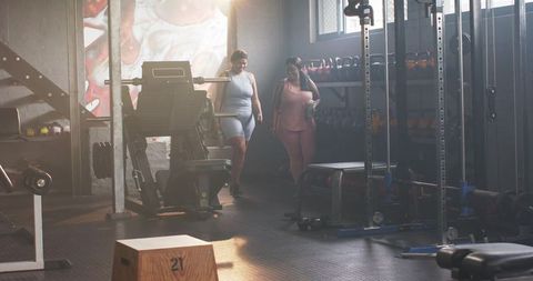 Diverse women walking through sunlit gym sharing post-workout camaraderie near weights