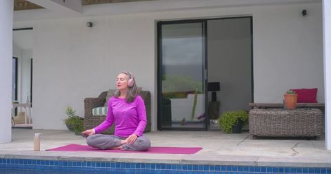 Middle-aged woman meditating poolside on pink mat wearing headphones for mindfulness