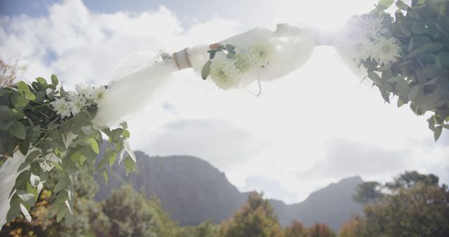 Decorated wedding arch with white flowers and draped fabric under sun in a picturesque mountain setting. Ideal for content on weddings, outdoor ceremonies, nature, romantic events, and scenic backdrops. Useful for wedding planning websites, event management blogs, and content about natural outdoor events aesthetics.