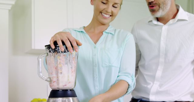 A smiling couple in a modern kitchen making a smoothie using a blender. This image can be used for advertisements related to healthy lifestyles, kitchen appliances, home cooking, and meal preparation. It is perfect for promoting family or couple-related products and activities.