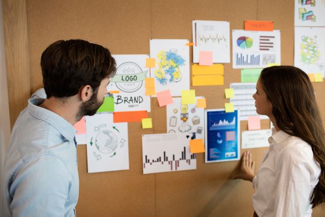 Business colleagues are analyzing various charts and graphs pinned on a soft board in an office. This image is ideal for illustrating teamwork, corporate planning, and collaborative brainstorming sessions. It can be used in business presentations, marketing materials, and articles related to business strategy and project management.