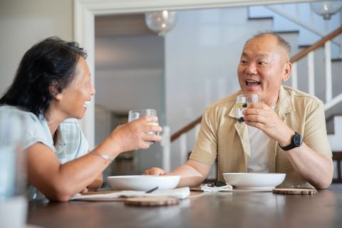 Asian senior couple toasting citrus drinks at home dining table sharing conversation