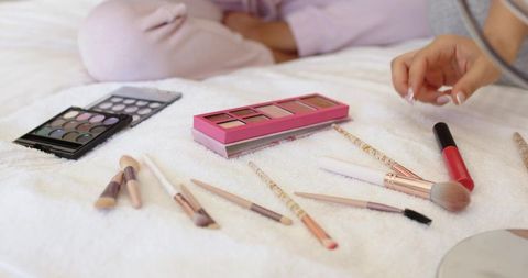 Friends arranging makeup brushes and palettes on bed in pale pink loungewear