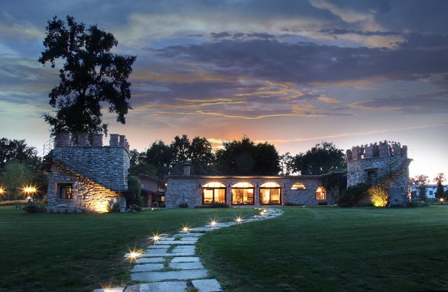 Luxuriously designed stone house with two towers surrounded by lush greenery. Evening setting with illuminated pathway leading to the home. Ideal for real estate listings, lifestyle magazines, or architectural design features highlighting luxury living, countryside properties, or architectural brilliance.