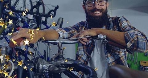 Smiling bearded mechanic leaning on bike handlebars in workshop with gold star confetti