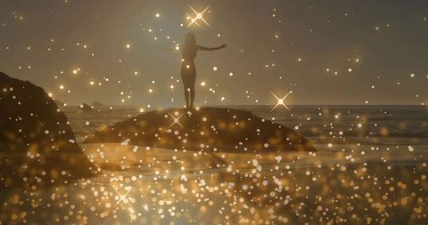 Standing woman raising arms on rocky outcrop at sunset with golden bokeh sparkle overlay