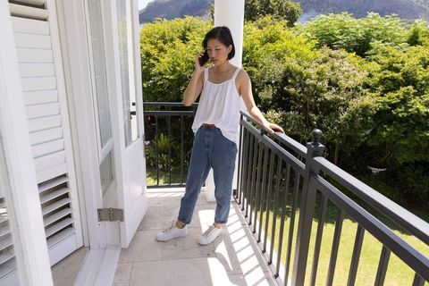 Asian woman standing on balcony using smartphone, casual summer outfit, garden sunlight