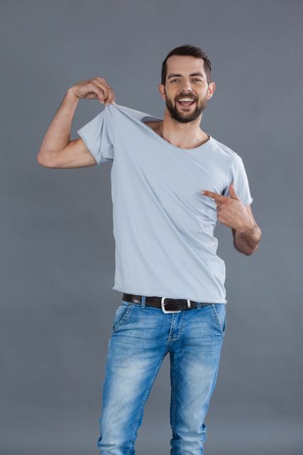 This image features a cheerful man posing confidently in a grey t-shirt and jeans against a grey background. Ideal for use in fashion, lifestyle, and casual wear promotions. Perfect for advertisements, social media content, and blog posts focusing on men's fashion and modern lifestyle.