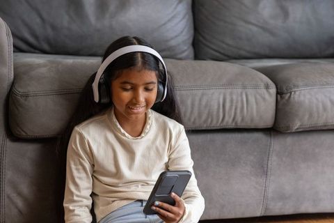 Young indian girl wearing over-ear headphones using smartphone while sitting on gray sofa