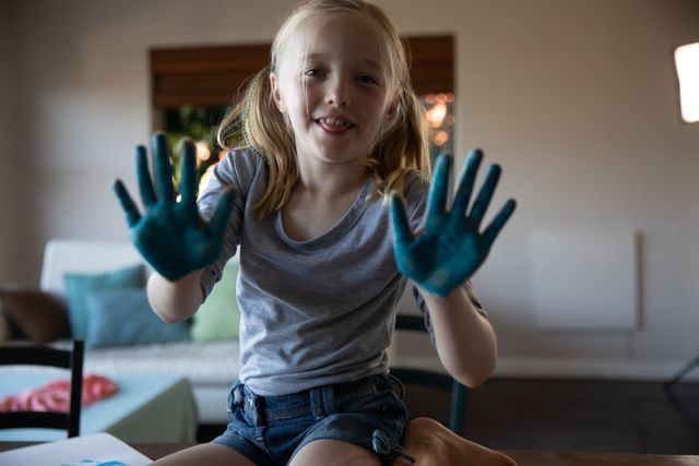 Portrait of Caucasian girl spending time at home, smiling to camera showing her hands painted blue doing creative art. Childhood leisure time at home.
