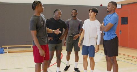Diverse male athletes standing and talking on indoor gym court with coach and whistle