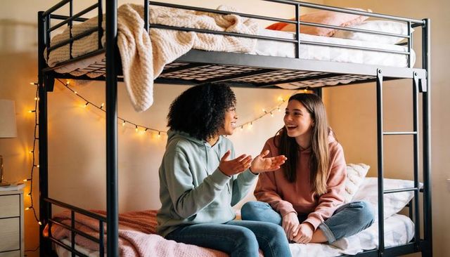Cozy dorm bunk chatting teen friends in hoodies and jeans with warm string lights