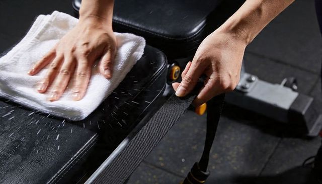 Hands wiping gym bench with white towel and gripping resistance band for equipment hygiene
