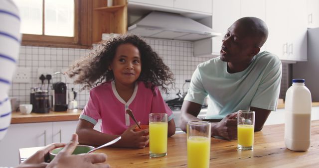 Father and daughter sitting at kitchen table enjoying breakfast together with glasses of orange juice. The atmosphere is casual and cheerful, emphasizing family bonding and togetherness. Useful for advertisements, parenting articles, and content related to family life and morning routines.
