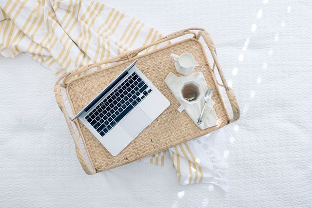 Photograph shows a wicker tray on a white bedspread with a laptop, a cup of tea, and a milk jug. This image can be used to suggest a peaceful morning routine, work-from-home scenarios, and achieving work-life balance. Ideal for lifestyle blogs, morning routine articles, work-from-home set-ups, and cozy living promotions.