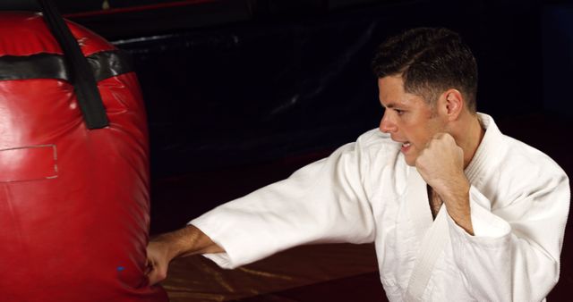 Male martial artist wearing white uniform, punching red heavy bag in gym. Used for fitness, sports training, martial arts techniques, determination, and discipline related themes.