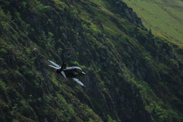 Military jet flying low over lush, green, mountainous terrain offers dramatic aviation scene. Useful for articles and ads themed around piloting, military exercises, environmental cinematography, and adventurous outdoor careers.