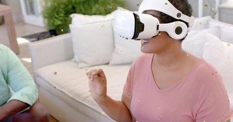 Smiling woman wearing white VR headset reaching for virtual stars on living room sofa